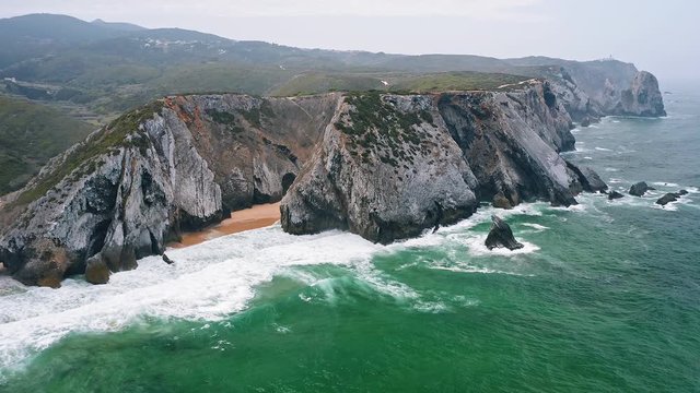 Aerial view of rocky cliff formation of Praia da Adraga beach, Portugal Atlantic coast. Cabo do Paco Lighthouse in Background