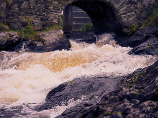 Water flows in a small river over a stone creating splash in focus, old bridge and river out of focus.