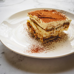 Close up of a slice of homemade tiramisu (traditional Italian dessert) on a white plate with a spoon on a marble board. Square format.
