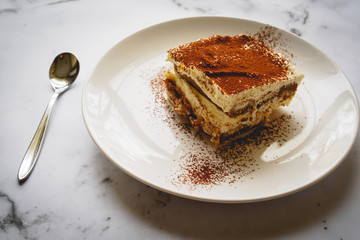 Close up of a slice of homemade tiramisu (traditional Italian dessert) on a white plate with a spoon on a marble board. Landscape format.