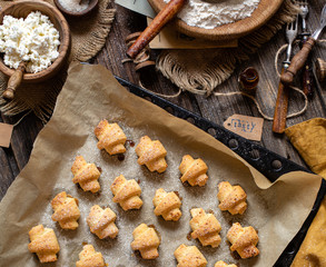 overhead shot of tasty baked rolled or bagels cookies on baking tray with parchment on rustic wooden table with bowls of flour, sugar and cottage cheese, orange napkin