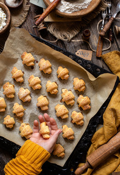 overhead shot of tasty baked rolled or bagels cookies on baking tray with parchment in woman hands on rustic wooden table with bowls of flour, sugar and cottage cheese, orange napkin