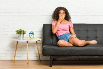 Young african american woman sitting on the sofa thoughtful looking to a copy space covering mouth with hand.