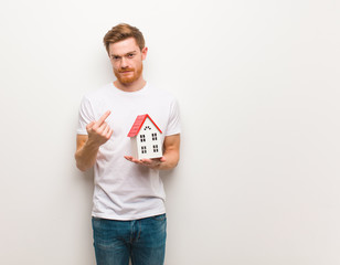 Young redhead man inviting to come. Holding a house model.