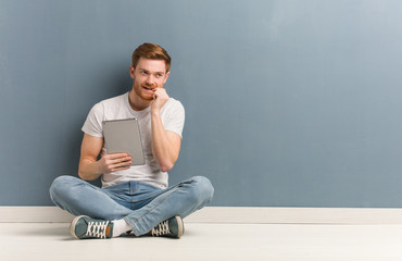 Young redhead student man sitting on the floor relaxed thinking about something looking at a copy space. He is holding a tablet.