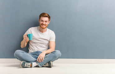 Young redhead student man sitting on the floor cheerful with a big smile. He is holding a coffee mug.