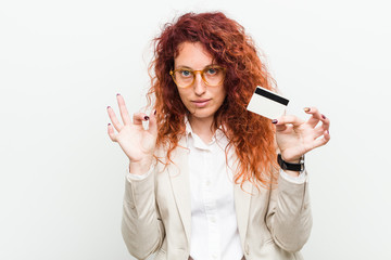 Young caucasian redhead woman holding a credit card cheerful and confident showing ok gesture.