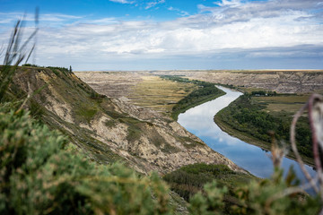 Reflective river cutting through the Alberta Badlands with exposed sedimentary rock canyon and shrubs  - obrazy, fototapety, plakaty