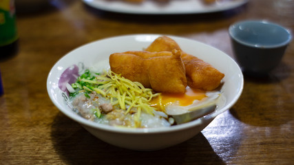Asian congee with minced pork, sliced ginger, vegetable and mini deep-fried dough sticks in glass bowl on wooden table. a delicious traditional Asian food style