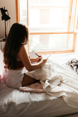 Obraz na płótnie Canvas Young brunette woman writing in a notebook on bed at home in the morning. Girl enjoying novel in bedroom, education lifestyle concept. Young woman reading book on bed at home