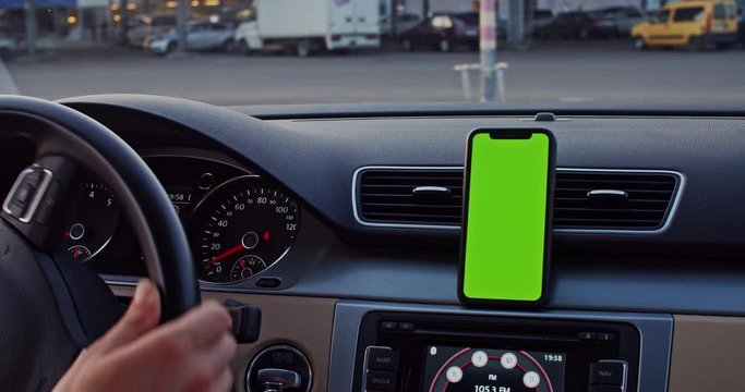 Person driving car in shopping mall parking space using mobile app on blank greenscreen mock-up smartphone on dashboard close-up.