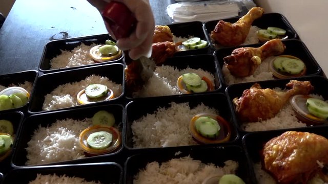 A row of Nasi Lemak in lunch boxes container while hands put a chicken roasted on it.