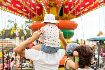 Child sitting on her father's shoulders in front of a colorful carousel at a carnival. Girl and boy with their parents at the vintage amusement park rides. Wearing a cute ladybug outfit and a sun hat. © studiolaska