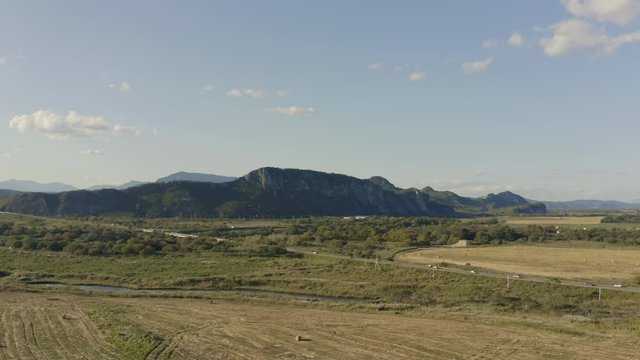 Landscape aerial shot of the countryside road with moving cars passing through the fields with haystacks and mountain ridge in the background on a clear sunny day, Russia, far East.