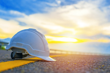 Fototapeta na wymiar Close up of white safety helmet placed on the yellow solid line of road with sunset and blue sky background, Helmet in construction site after work during the sunset, Safety and Engineering Concept.