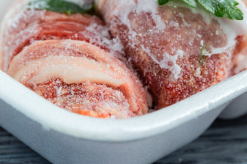 Frozen meat in hoarfrost in a white plastic tray with greens.