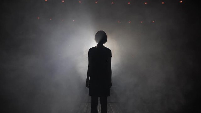 dark silhouette of young woman with short haircut looking at bright spotlight creating halo in studio slow motion