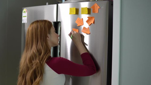 A young beautiful young European model with long Reddish Brown hair writes her to do list on post it notes on a stainless steel fridge. She is planning her day with positive affirmations