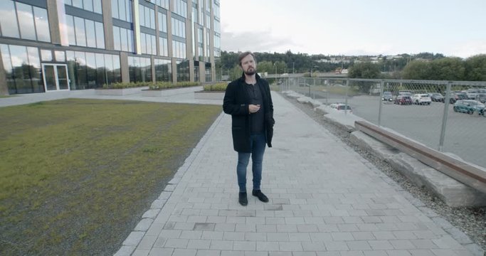 Young man with beard in black wool coat standing in a parking lot eating granola bar camera pusing in