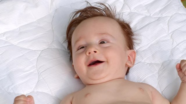 White smiling little child with dark thick hair and dark eyes is lying topless on the blanket on open air and observes the area. It is moving head and fingers. It raises left and right hand