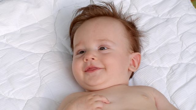 White smiling little child with dark thick hair and dark eyes is lying topless on the blanket on open air and observes the area. It is moving head and fingers. It raises left and right hand