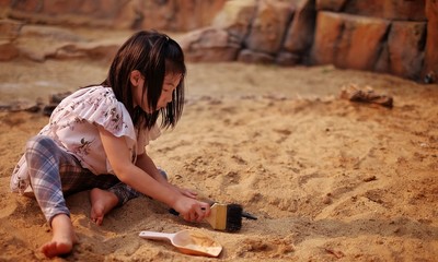 A young Asian girl playing in a sandbox with a modeled dinosaur fossil using brush and shovel.