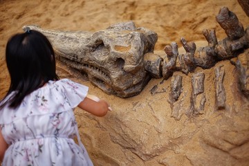 A girl playing in a sandbox with a modeled dinosaur fossil, digging sand off the fossil.