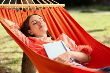 Beautiful young woman with book resting in hammock outdoors