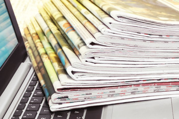 Newspapers and laptop. Pile of daily papers with news on the computer. Pages with headlines, articles folded and stacked on keypad of electronic device. Modern gadget and old journals, focus on paper 
