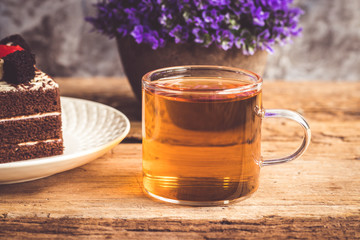 Hot tea In clear glass, black forest creek cake decorate with cherry and cake brownies In a white plate, and have false heather In pots small, all place are on wooden table.