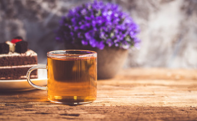 Hot tea In clear glass, black forest creek cake decorate with cherry and cake brownies In a white plate, and have false heather In pots small, all place are on wooden table.