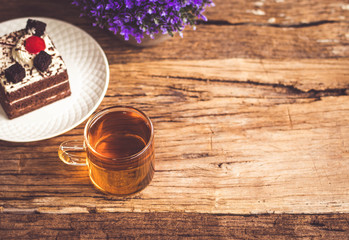 Fototapeta na wymiar Hot tea In clear glass, black forest creek cake decorate with cherry and cake brownies In a white plate, and have false heather In pots small, all place are on wooden table.