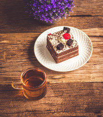 Hot tea In clear glass, black forest creek cake decorate with cherry and cake brownies In a white plate, and have false heather In pots small, all place are on wooden table.