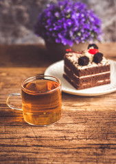 Hot tea In clear glass, black forest creek cake decorate with cherry and cake brownies In a white plate, and have false heather In pots small, all place are on wooden table.
