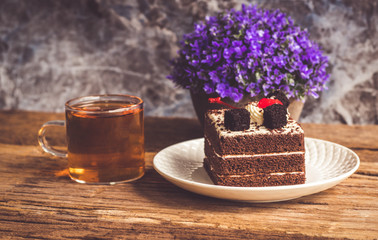 Hot tea In clear glass, black forest creek cake decorate with cherry and cake brownies In a white plate, and have false heather In pots small, all place are on wooden table.