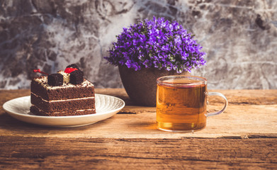 Hot tea In clear glass, black forest creek cake decorate with cherry and cake brownies In a white plate, and have false heather In pots small, all place are on wooden table.