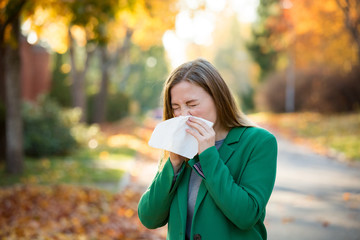 Sick young woman with cold and flu standing outdoors, sneezing, wiping nose with handkerchief,...