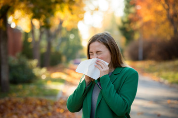 Sick young woman with cold and flu standing outdoors, sneezing, wiping nose with handkerchief,...