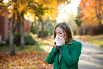 Sick young woman with cold and flu standing outdoors, sneezing, wiping nose with handkerchief, coughing. Autumn street background - obrazy, fototapety, plakaty