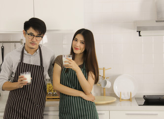 Young Asian couple standing in the kitchen holding glasses of milk for their breakfast and looking to the camera. copy space.
