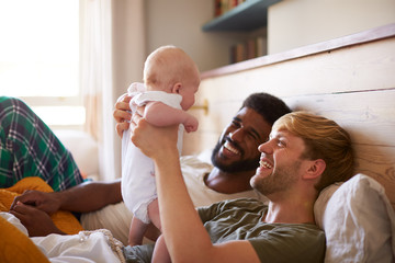 Loving Male Same Sex Couple Cuddling Baby Daughter In Bedroom At Home Together - obrazy, fototapety, plakaty