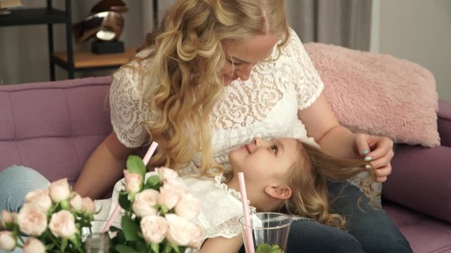 Portrait of young mother sits on couch and stroks blonde curly hair of 5 year old daughter, who lies on knees, look at each other, talk and smile. Concept of mother's love and care about child