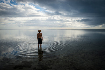 Tropical silhouette of man in straw hat standing in calm shallow waters with ripples emanating on...