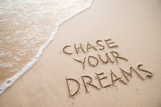 Chase Your Dreams travel message handwritten on smooth sand beach with incoming tropical wave