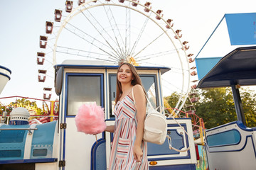 Charming young brunette lady with long brown hair posing over ferris wheel on sunny warm day, wearing romantic dress and white backpack, holding cotton candy on stick