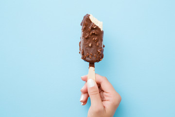Young woman hand holding white vanilla ice cream with nuts and chocolate glaze on pastel light blue background. Bitten food. Closeup.