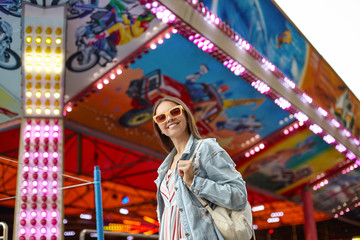 Obraz na płótnie Canvas Outdoor shot of beautiful young female in sunglasses walking through amusement park on warm day, wearing romantic dress and jeans coat, smiling sincerely to camera