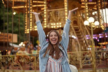 Happy young brunette lady with long hair standing over carousel in park of attractions, raising hands joyfully with wide mouth opened, positive emotions concept