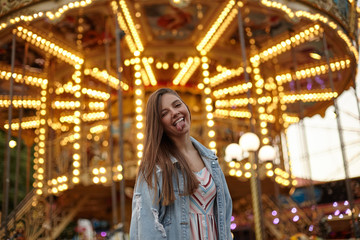 Funny outdoor photo of beautiful young woman with long hair, posing over attractions in amusement park, giving wink to camera and showing tongue