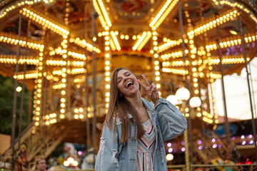 Joyful young attractive woman with brown long hair posing over carousel in jeans coat, raising hand with victory gesture, smiling with wide mouth opened and closing eyes